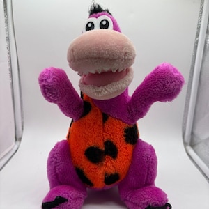 May include: A plush purple dinosaur toy with a pink snout, white teeth, and large eyes. The dinosaur has an orange belly with black spots and is posed with its arms up. The toy has black claws and a small tuft of black hair.