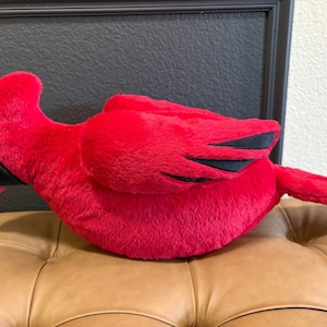 May include: A plush, bright red cardinal bird with black accents on the wings and face. The stuffed animal is lying on a brown leather surface, with a black frame in the background.
