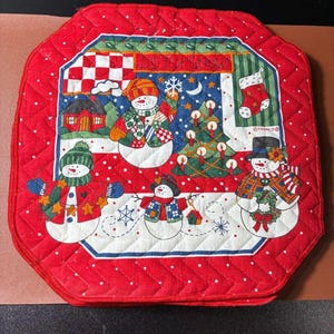 May include: Red quilted Christmas trivet with a snowman design. The trivet features a central panel with snowmen, a Christmas tree, and the words "LET IT SNOW". The border is red with white polka dots.