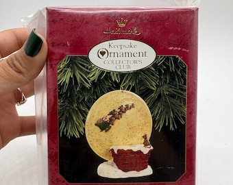 May include: A Hallmark Keepsake Ornament Collector's Club box. The red box features a Christmas ornament with a chimney, moon, and Santa's sleigh. The text on the box reads "Happy Christmas to All!" and "1997 Keepsake of Membership."