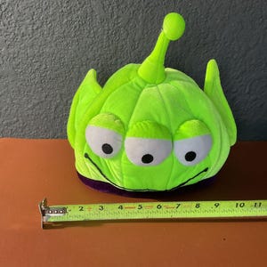 Pode incluir: Um chapéu de pelúcia verde limão, com três olhos com bordas brancas e pupilas pretas. O chapéu tem uma faixa roxa na base e uma única antena. O chapéu mede aproximadamente 25 cm de comprimento.