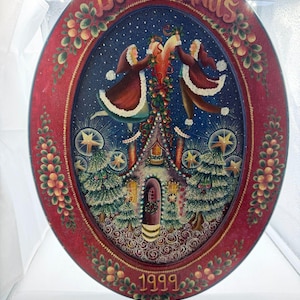 May include: An oval decorative wooden plaque featuring a hand-painted winter scene with two figures holding a heart above a house, surrounded by pine trees and stars. The red border displays the text DECK the HALLS and the year 1999.