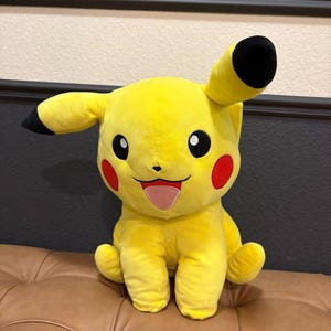 May include: A plush Pikachu toy, featuring bright yellow fur, black-tipped ears, and red cheek circles. The toy has a smiling expression with a pink tongue and is sitting on a brown surface.