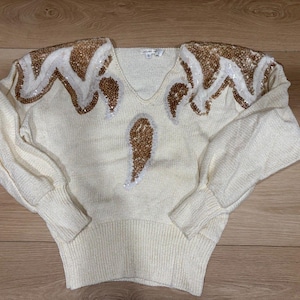 Pode incluir: Camisola creme com decote em V e mangas compridas. Os ombros e o centro da camisola são adornados com desenhos de lantejoulas douradas e brancas. A camisola é feita de um material de malha macia.