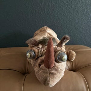 May include: A tan plush rhinoceros toy featuring a single dark brown fabric horn, textured skin-like fabric, and two large, protruding plastic eyes with visible pupils and irises, positioned on either side of the central horn on the head.