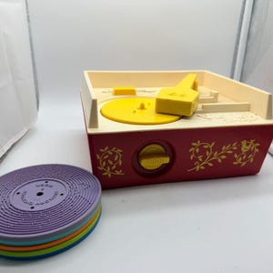 May include: A vintage toy record player with a cream-colored top and a maroon base featuring gold vine and bird illustrations. It includes a yellow turntable, a yellow tone arm, and a stack of colorful plastic discs in purple, orange, green, and blue.