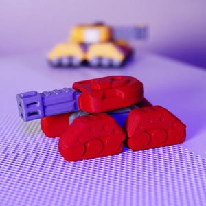 May include: A red and purple toy tank with a detailed design. The tank has a red body, a purple cannon, and red treads. Another toy tank is visible in the background, with yellow and purple colors.
