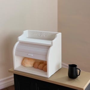 May include: A white miniature breadbox with a roll-top door, holding several small, round loaves of bread. A small black pitcher sits on the counter next to the breadbox. The breadbox has a decorative floral design on the top.