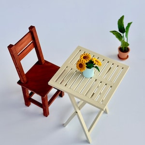 Può includere: Un set da patio in miniatura in legno con una sedia rossa, un tavolo color crema e una piccola pianta in vaso. Il tavolo ha un vaso azzurro con girasoli gialli. Il set è progettato per una casa delle bambole o una scena in miniatura.
