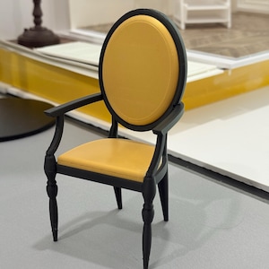 Puede incluir: Una silla negra en miniatura con respaldo y asiento ovalados amarillos. La silla tiene reposabrazos curvos y patas torneadas. La silla es un modelo a pequeña escala, probablemente para una casa de muñecas o una exhibición.