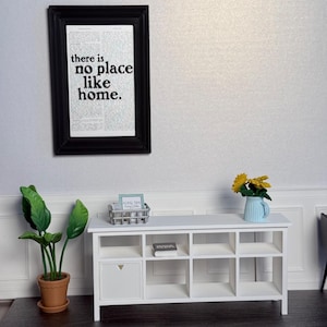 Może przedstawiać: Miniaturowa biała szafka z otwartymi półkami i zamkniętymi drzwiami szafki. Oprawiony nad szafką napis brzmi "there is no place like home". Obecna jest również roślina doniczkowa i niebieski dzbanek z żółtymi kwiatami.