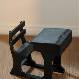 May include: A small black school desk and chair. The desk has a hinged top and is attached to the chair. The chair has a slatted back. The set is made of a dark material and is on a wooden surface.