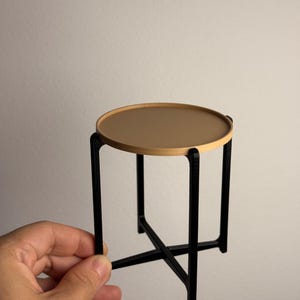 May include: A miniature round table with a light brown top and black legs. The table's top is approximately 4 inches in diameter, supported by a black, geometric base. The table is held by a hand.