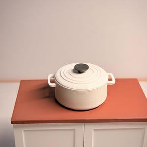 Könnte beinhalten: Ein weißer emaillierter gusseiserner Bräter mit schwarzem Knopf und seitlichen Griffen. Der Topf steht auf einer rot-orangen Arbeitsplatte. Das Kochgeschirr steht auf einem weißen Schrank.