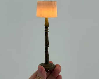 Può includere: Una lampada da tavolo in miniatura con base e stelo marroni, sormontata da un paralume a coste color crema. La lampada è accesa, emettendo un bagliore caldo e invitante. La lampada è tenuta in mano su uno sfondo bianco.
