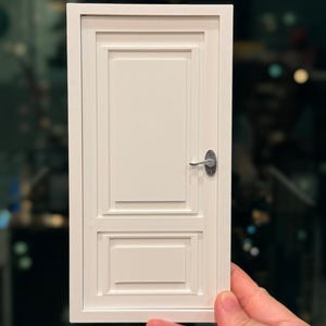 Könnte beinhalten: Eine kleine, weiße Tür mit einem silbernen Griff. Die Tür hat einen rechteckigen Rahmen und Paneeldetails. Die Tür wird von einer Hand vor einem verschwommenen Hintergrund gehalten.