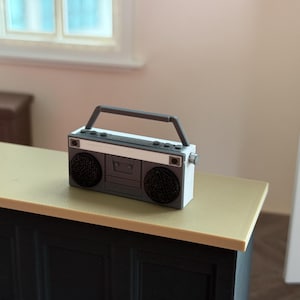 Könnte beinhalten: Ein Miniatur-Boombox im Retro-Stil in Grau-, Weiß- und Schwarztönen mit einem silbernen Griff. Das kleine Radio steht auf einer hellgelben Oberfläche, mit einem Fenster und einer Tür im Hintergrund.