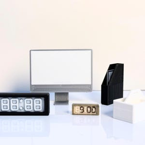 Könnte beinhalten: Eine Miniatur-Büroszene mit einem silbernen Computermonitor, einem schwarzen Zeitschriftenhalter, einer weißen Box und zwei Digitaluhren, die "88:88" und "09:00" anzeigen. Die Objekte sind auf einer reflektierenden Oberfläche angeordnet.