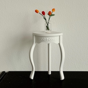Könnte beinhalten: Ein kleiner, weißer Beistelltisch mit drei Beinen und einem dekorativen Blumendesign. Eine kleine Glasvase enthält orangefarbene und rote Blumen. Der Tisch steht vor einer weißen Wand. Der Tisch ist etwa 40 cm hoch.