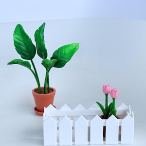 May include: A miniature scene featuring potted plants. A green-leafed plant and pink tulips are in terracotta pots. A small white picket fence surrounds the tulips. The scene is designed to be decorative and charming.