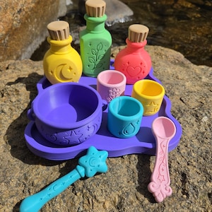 May include: A set of colorful plastic potion-making toys including a purple tray, a purple cauldron, three bottles with tan corks, two small cups, a light blue star-topped wand, and a pink spoon, all featuring embossed symbols like moons, stars, and hearts.