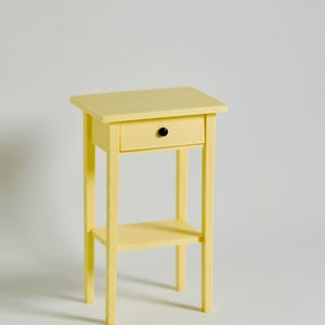 Puede incluir: Una mesita de noche pequeña y amarilla con un solo cajón y un estante inferior. La mesita de noche tiene un pomo negro en el cajón y está pintada en un color amarillo pálido liso. La mesa mide aproximadamente 61 cm de altura.