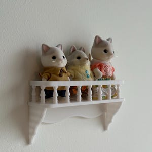 May include: Three cat figurines on a white decorative shelf. The cats wear various outfits, including a yellow coat, a pink top, and a mustard-colored jacket. The shelf is mounted on a white wall.