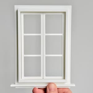 Op de afbeelding: Een klein wit raam met zes ruiten, een vensterbank en een frame. Het raam wordt tegen een grijze achtergrond gehouden. Het raam is een decoratief item.