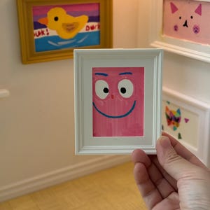 May include: A small framed artwork featuring a pink face with a blue smile and white eyes. The painting is held in front of a wall with other framed art, including a yellow duck painting and a pink cat painting.