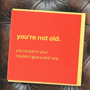 May include: A red greeting card with yellow borders and the text "you're not old. you're just in your 'couldn't give a shit' era." The card is on a brown, textured surface.