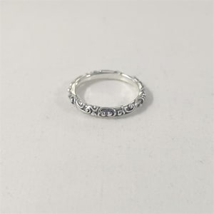 May include: A silver ring with an intricate, repeating scrollwork design. The ring is thin and appears to be made of metal. The detailed pattern covers the entire band, creating a delicate and ornate appearance. The ring is shown on a white surface.