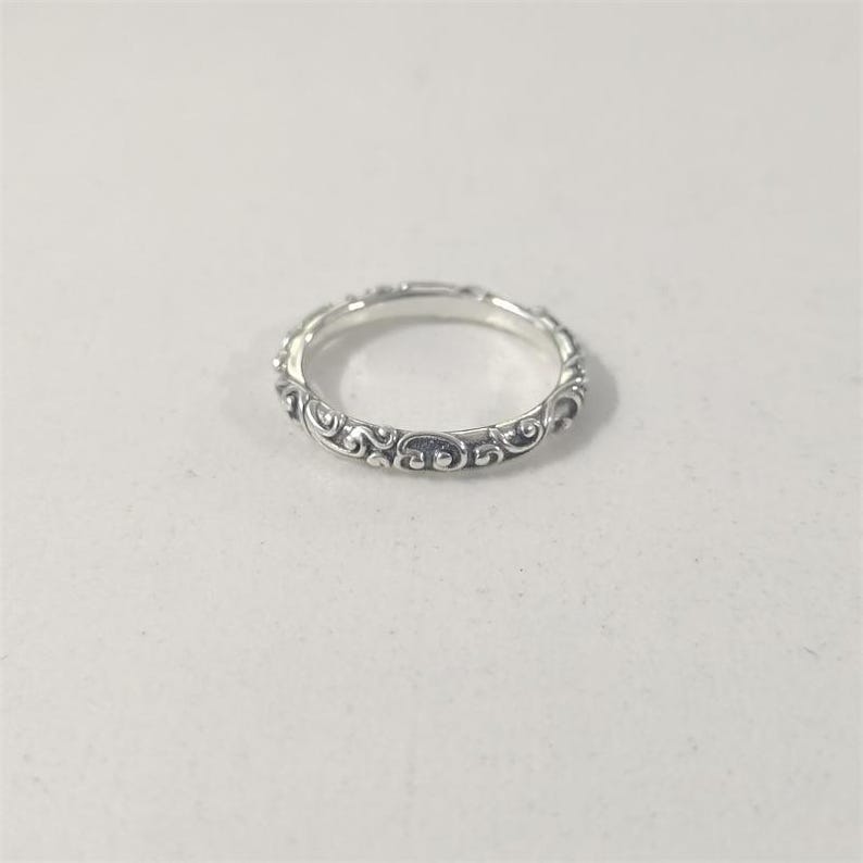 May include: A silver ring with an intricate, repeating scrollwork design. The ring is thin and appears to be made of metal. The detailed pattern covers the entire band, creating a delicate and ornate appearance. The ring is shown on a white surface.