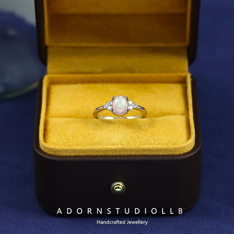 May include: A silver ring featuring an oval opal gemstone and smaller accent stones, presented in a brown jewellery box with a yellow interior. The ring is set against a blue backdrop. The text "ADORNSTUDIOLLB Handcrafted Jewellery" is visible.