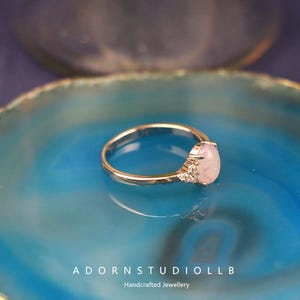 May include: A gold ring featuring a pear-shaped, pale pink gemstone. The ring is displayed on a blue surface. The image includes the text "ADORNSTUDIOLL B" and "Handcrafted Jewellery" at the bottom.