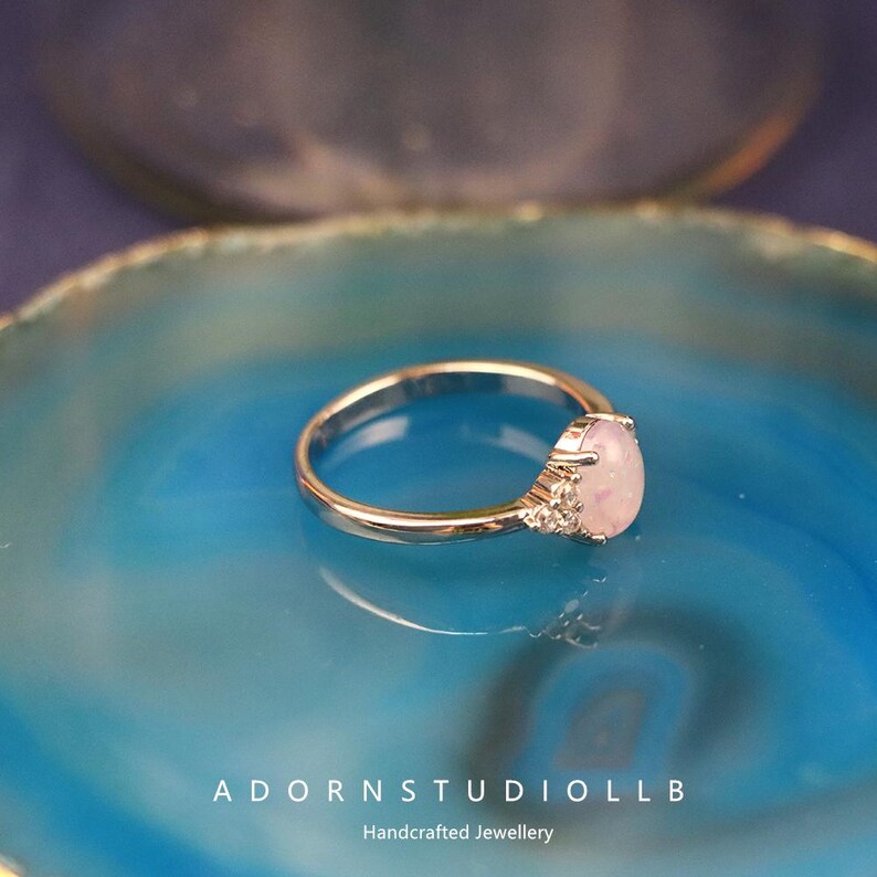 May include: A gold ring featuring a pear-shaped, pale pink gemstone. The ring is displayed on a blue surface. The image includes the text "ADORNSTUDIOLL B" and "Handcrafted Jewellery" at the bottom.