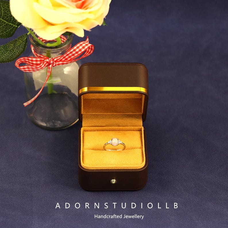 May include: A brown jewelry box with gold trim, open to display a ring with an opal and two smaller stones. The box is on a blue surface, beside a vase with a yellow rose and a red and white ribbon. The text "ADORNSTUDIOLLB" and "Handcrafted Jewellery" are at the bottom.