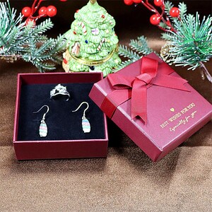 May include: A red gift box with a matching ribbon and the text "Best Wishes For You, Especially for you". Inside, a ring and a pair of earrings are displayed in a red box with a black interior. A small Christmas tree is in the background.