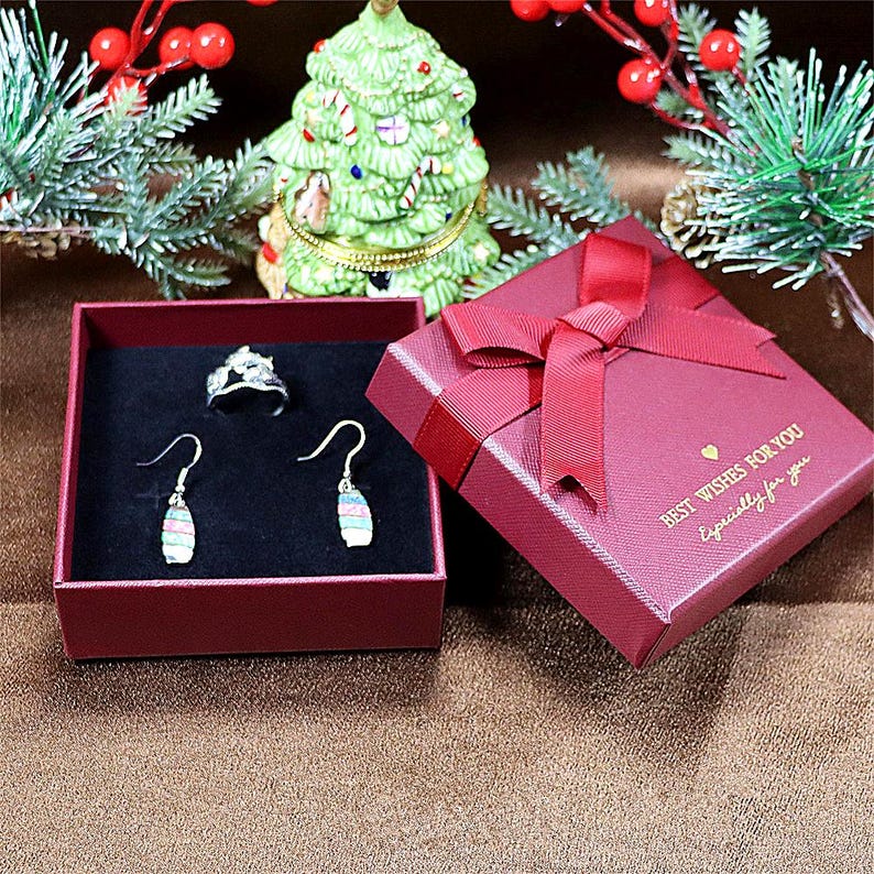May include: A red gift box with a matching ribbon and the text "Best Wishes For You, Especially for you". Inside, a ring and a pair of earrings are displayed in a red box with a black interior. A small Christmas tree is in the background.