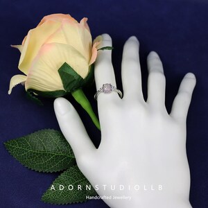 May include: A silver ring with a light purple gemstone is displayed on a white hand. The ring has a central stone and two smaller stones on either side. A yellow and pink rose and green leaves are in the background. The text "ADORNSTUDIOLLB Handcrafted Jewellery" is at the bottom.