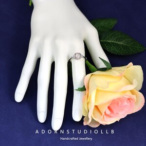 May include: A silver ring featuring a central oval, pale pink gemstone, with smaller clear stones either side. The ring is shown on a white hand, beside a yellow and pink rose. Text at the bottom reads "ADORNSTUDIOLLB Handcrafted Jewellery".