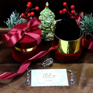 May include: A Christmas-themed display featuring a red velvet gift box and a gold-edged container, both with red ribbon. A silver ring and earrings with blue gemstones are shown with a thank you card. A small Christmas tree is in the background.