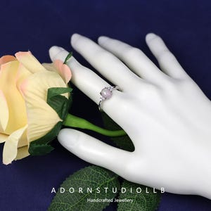 May include: A silver ring featuring a round, pale purple gemstone and small clear stones. The ring is displayed on a white hand, with a rose and the text "ADORNSTUDIOLLB Handcrafted Jewellery" in the background.