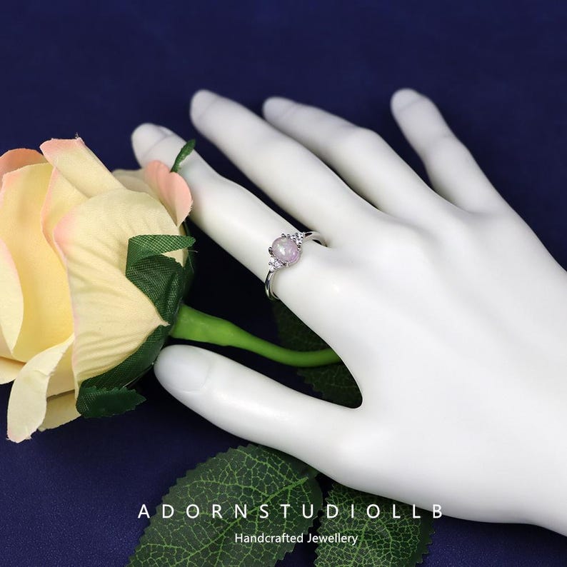 May include: A silver ring featuring a round, pale purple gemstone and small clear stones. The ring is displayed on a white hand, with a rose and the text "ADORNSTUDIOLLB Handcrafted Jewellery" in the background.