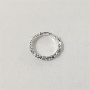 May include: A silver ring with an intricate, repeating scroll design. The ring is circular and sits on a white surface. The detailed pattern wraps around the entire band, creating a delicate and ornate appearance. The ring is likely made of sterling silver.