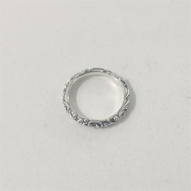 May include: A silver ring with an intricate, repeating scroll design. The ring is circular and sits on a white surface. The detailed pattern wraps around the entire band, creating a delicate and ornate appearance. The ring is likely made of sterling silver.