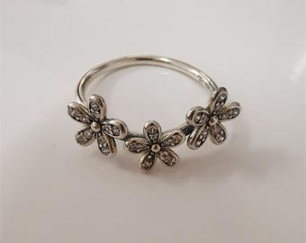 May include: A silver ring featuring three daisy flower designs. Each flower has a central stone and is surrounded by smaller, clear stones. The ring is set against a white background.