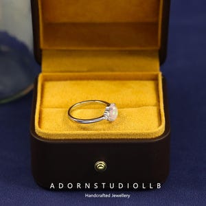 May include: A silver ring featuring an opalescent, pear-shaped gemstone, set in a fine band. The ring is presented in a brown jewellery box with a yellow velvet lining. The text "ADORNSTUDIOLL B Handcrafted Jewellery" is visible.