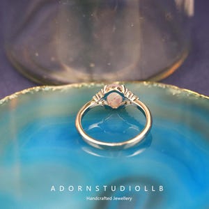 May include: A silver ring featuring a light pink oval gemstone, with smaller clear stones on either side. The ring is displayed on a blue and turquoise surface. The text "ADORNSTUDIOLLB Handcrafted Jewellery" is visible.