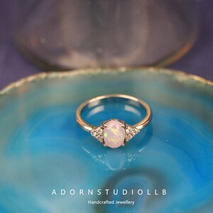May include: A silver ring featuring an oval opal gemstone and small accent stones. The ring is displayed on a blue surface. The text "ADORNSTUDIOLLB Handcrafted Jewellery" is visible at the bottom of the image.