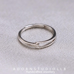 May include: A silver ring with a distinctive open design. The ring has a slim band with a rectangular cutout. It is presented on a light grey surface. The text "ADORNSTUDIOLL B Handcrafted Jewellery" is visible at the bottom.